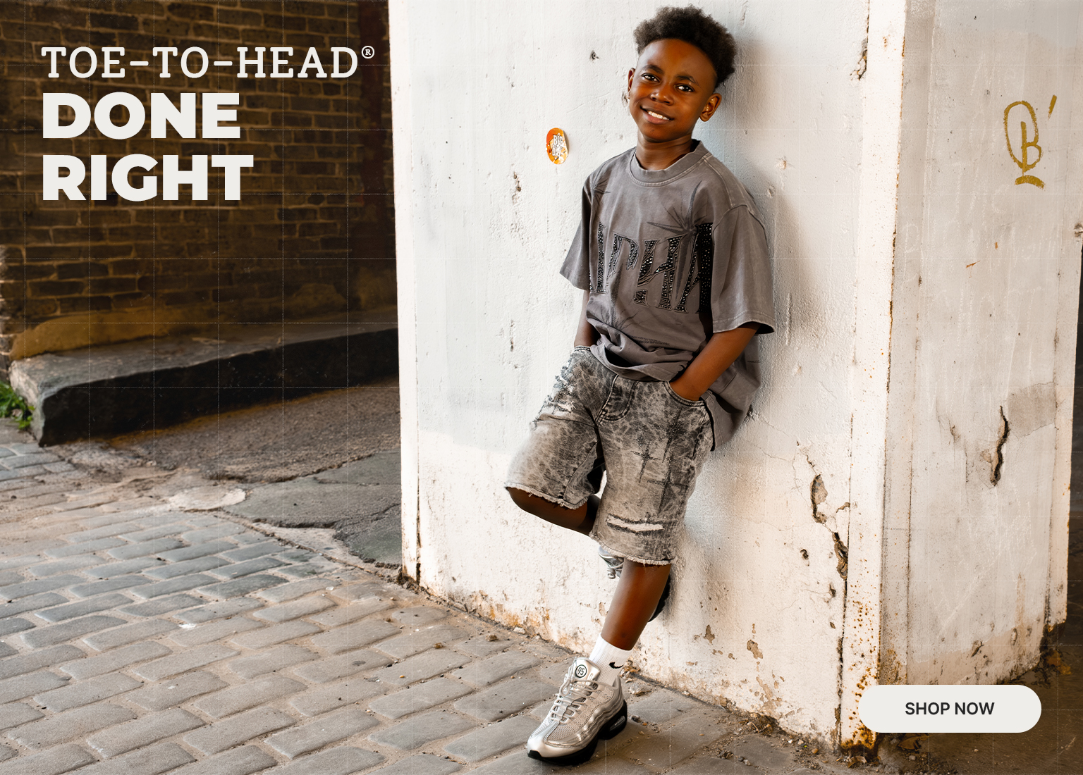 Promotional Hibbett banner featuring a child wearing a graphic T-shirt, denim shorts, crew socks, and athletic sneakers in an urban setting. The image includes the text “Toe-to-Head Done Right” and a “Shop Now” call-to-action, highlighting complete back-to-school apparel and footwear looks from Hibbett.